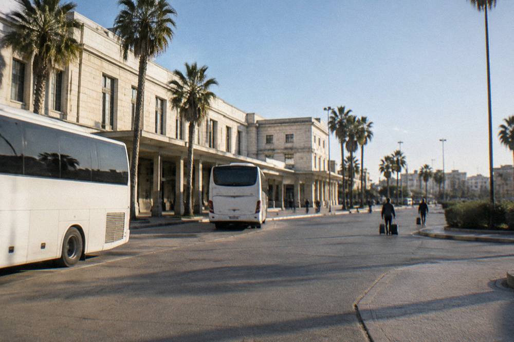 Station forecourt with plain white buses, palm trees and distant travellers with wheeled suitcases