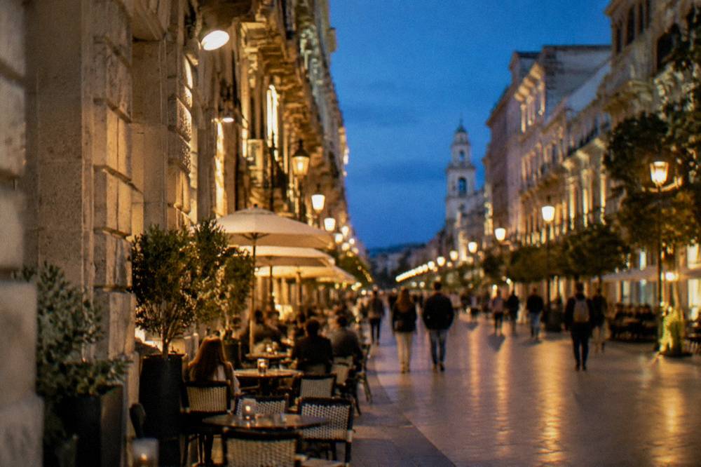 Pedestrian shopping street in the evening with lit lamps, cafe tables and out-of-focus people