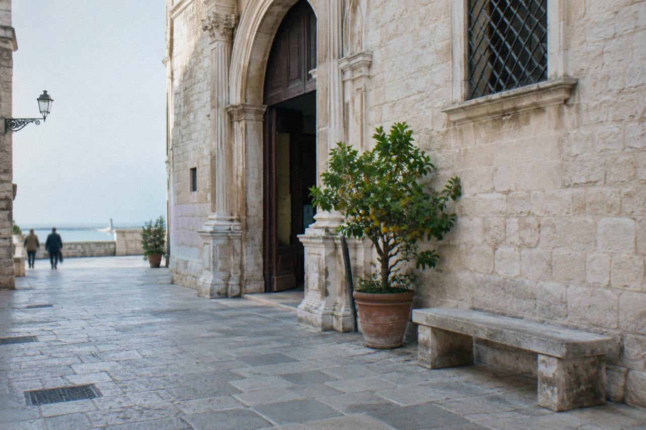 Pale stone palazzo with an arched portal on a quiet old-town street beside a stone bench and a potted lemon tree