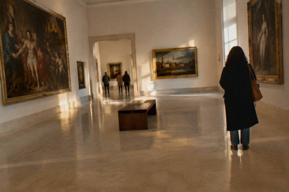 Gallery room with framed old paintings on pale walls, a central bench and one visitor seen from behind