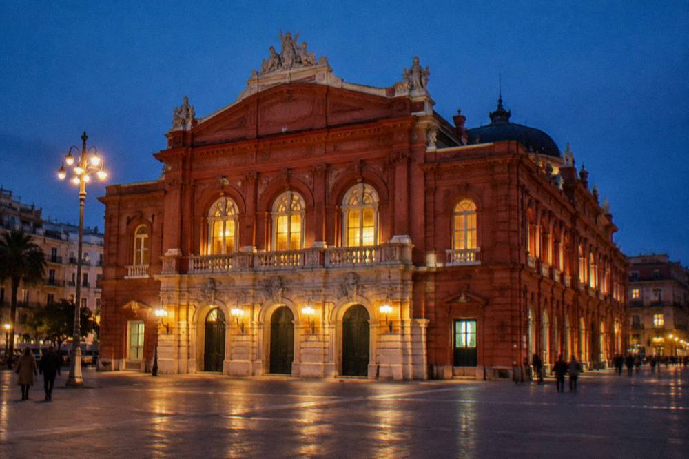 Neoclassical theatre facade floodlit at evening with glowing arched windows and a deep blue sky