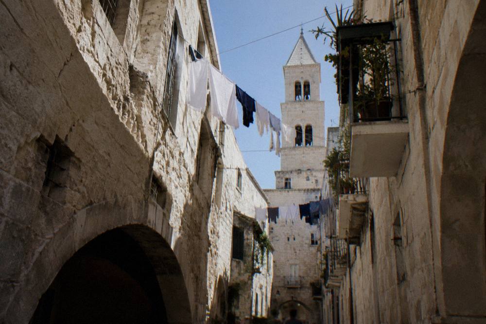 Looking up between close walls at a strip of blue sky crossed by washing lines and a bell tower
