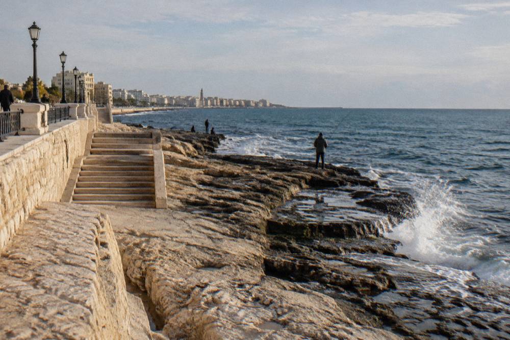Stone steps down to flat limestone rocks below a sea wall with distant anglers fishing and light sea spray