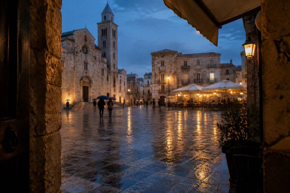 Rainy evening in a pale stone square with wet paving reflecting warm cafe lights and distant umbrellas