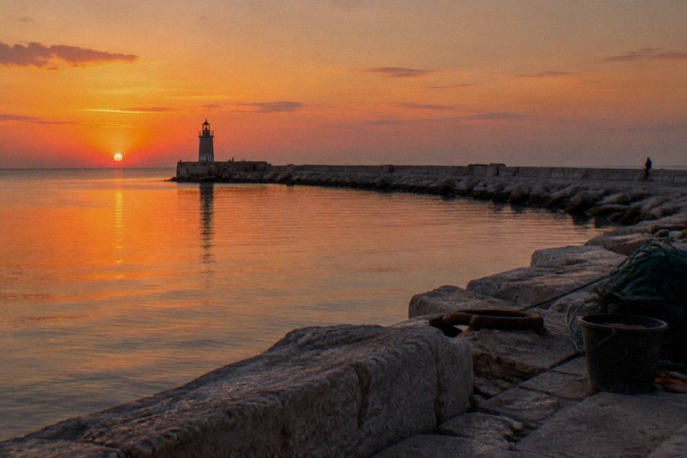End of a harbour pier with a small lighthouse, glassy open Adriatic and a distant angler at sunset