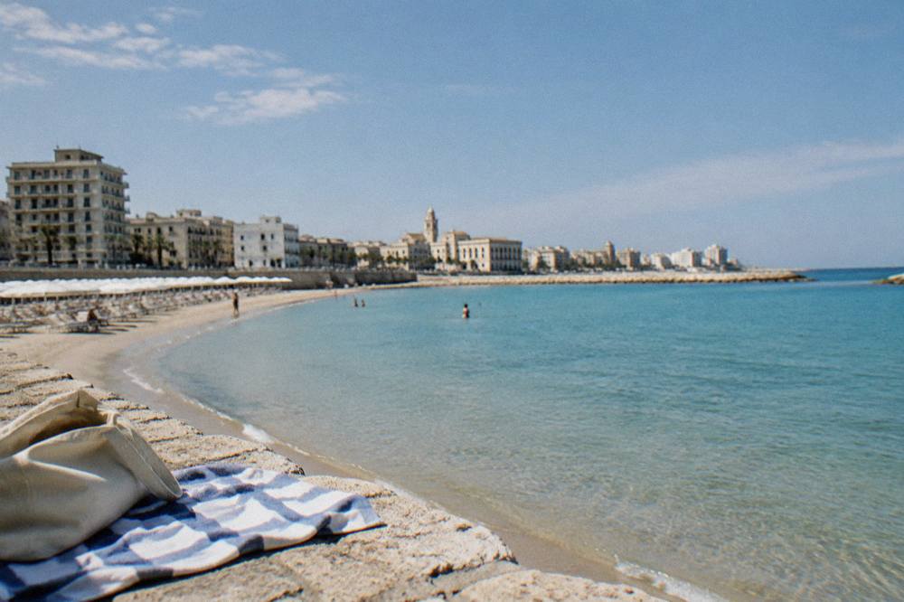 Small urban sandy beach with parasols in a curved bay, shallow turquoise water and seafront buildings behind