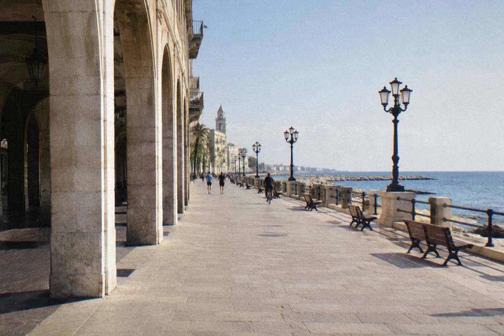Wide paved seafront promenade with distant joggers and cyclists, benches and calm blue sea in late afternoon