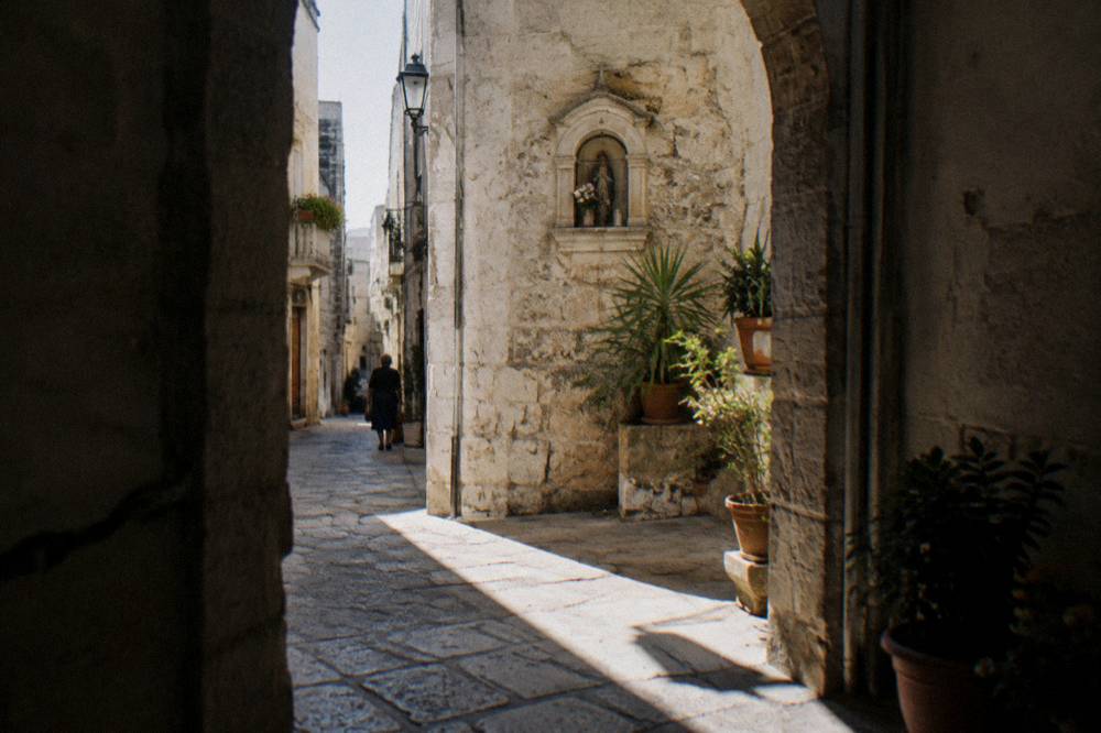 Crossing of narrow old town lanes with a stone shrine niche, potted plants and sunlight slicing between buildings