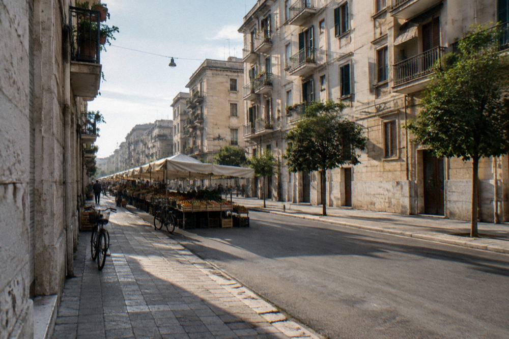 Residential street of apartment blocks with fruit and vegetable stalls under plain awnings and distant shoppers
