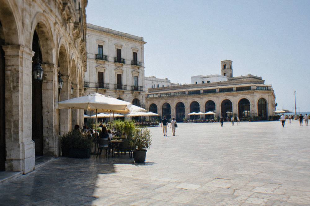 Broad open piazza beside a long low market hall with arched openings and outdoor cafe tables in bright morning