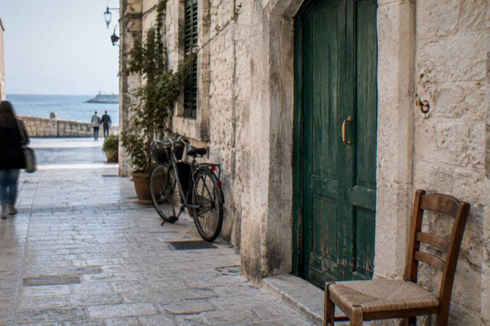 Faded green wooden door in a pale limestone wall with a chair and a bicycle beside it