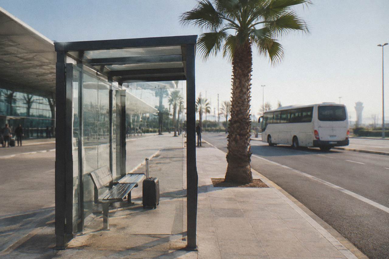 Bus shelter outside an airport terminal with a bench, a suitcase and a plain white bus waiting behind