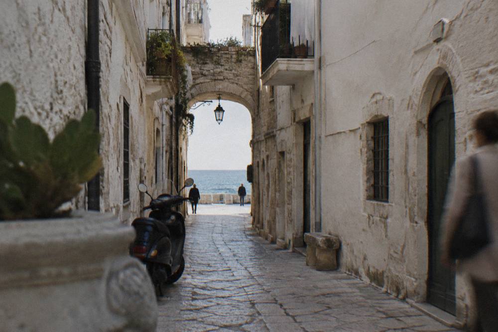 Straight old town lane between whitewashed walls ending in a bright slit of open sea with a parked scooter