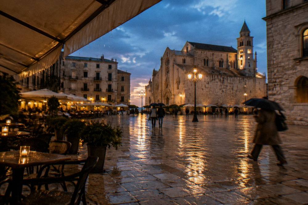 Rainy evening in a pale stone square with wet paving reflecting warm cafe lights and distant umbrellas