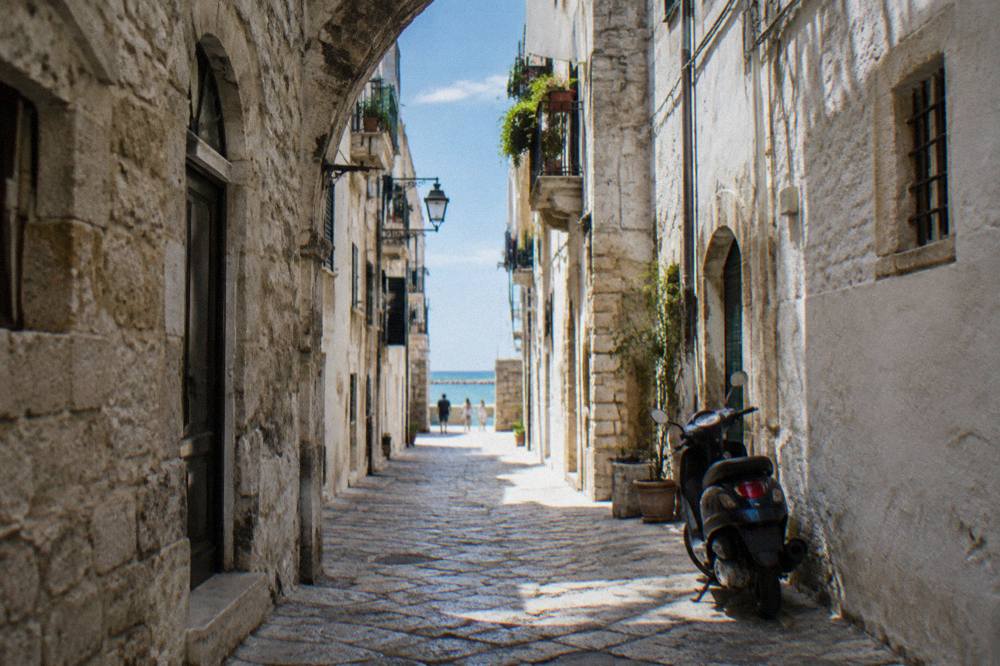 Straight old town lane between whitewashed walls ending in a bright slit of open sea with a parked scooter