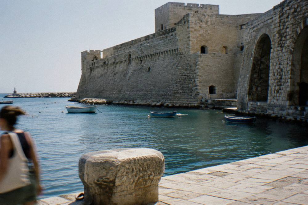Seaward wall of a pale stone castle beside harbour water with a few small boats in afternoon sun