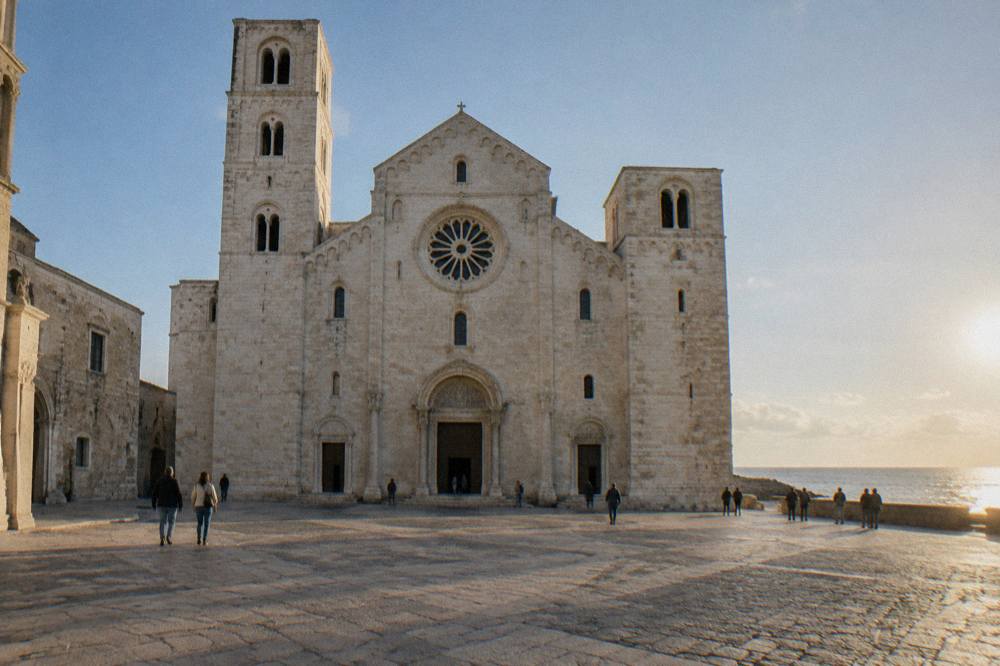 Romanesque church of pale limestone with a wide plain facade, two squat towers and a rose window above a paved square