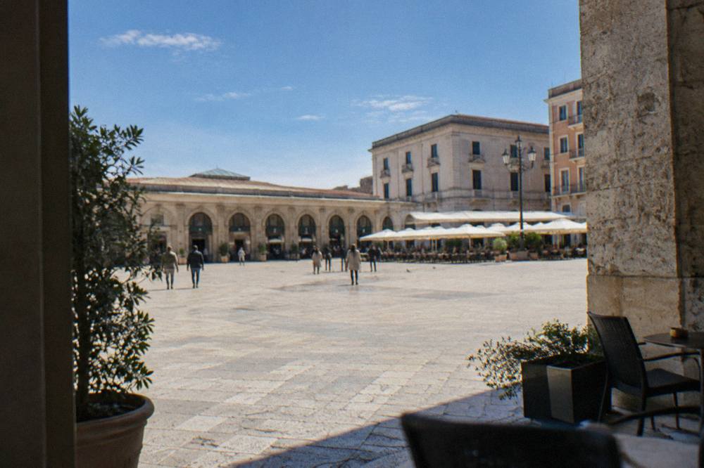 Broad open piazza beside a long low market hall with arched openings and outdoor cafe tables in bright morning