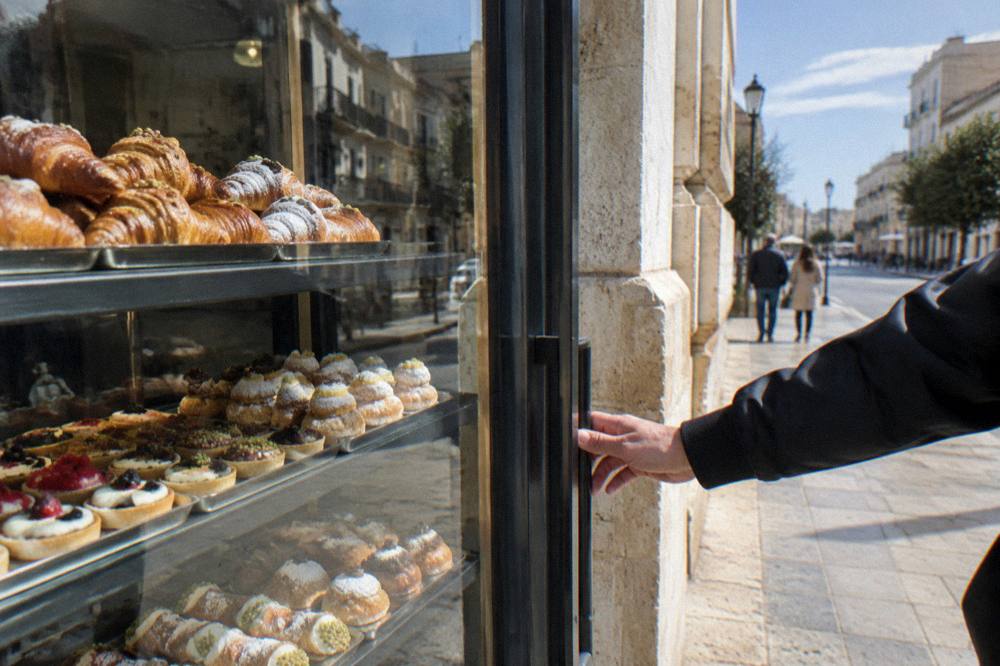 Pastry shop window with trays of pastries behind glass and a hand reaching for the door