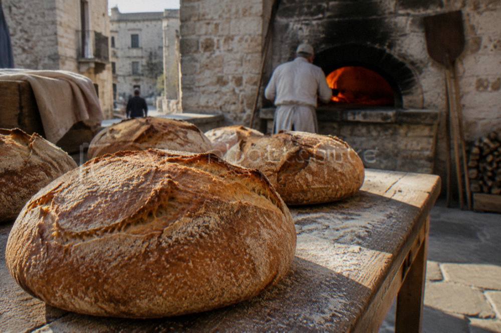 Large round loaves of semolina bread with thick golden crusts on a wooden table beside a wood-fired oven