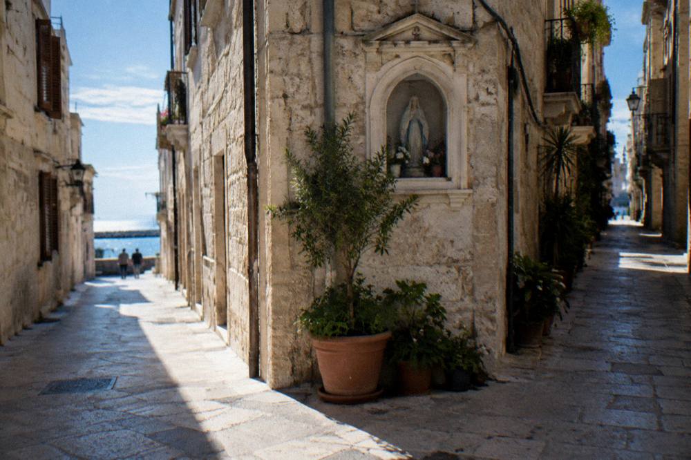 Crossing of narrow old town lanes with a stone shrine niche, potted plants and sunlight slicing between buildings