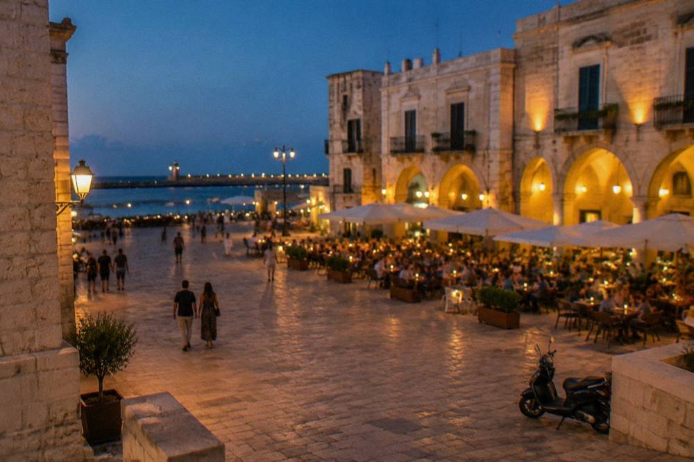 Evening piazza with warm light on cream walls, out-of-focus diners at tables and glowing market arches