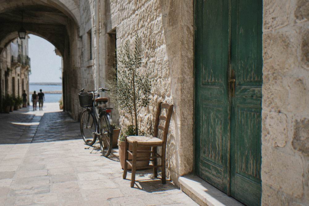 Faded green wooden door in a pale limestone wall with a chair and a bicycle beside it