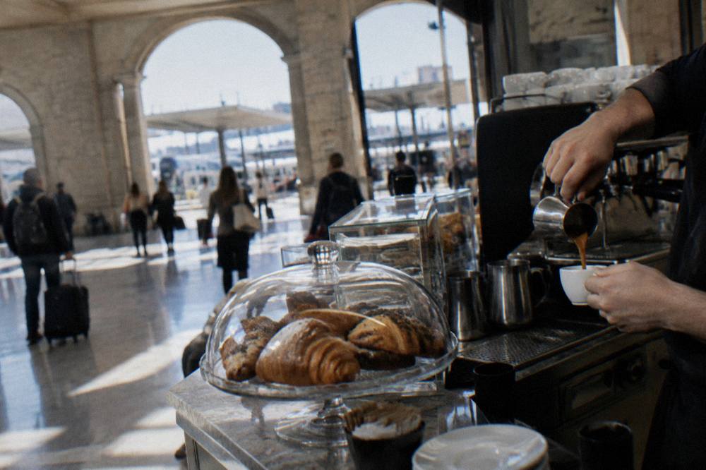 Station cafe counter with hands pouring an espresso and pastries under a glass dome