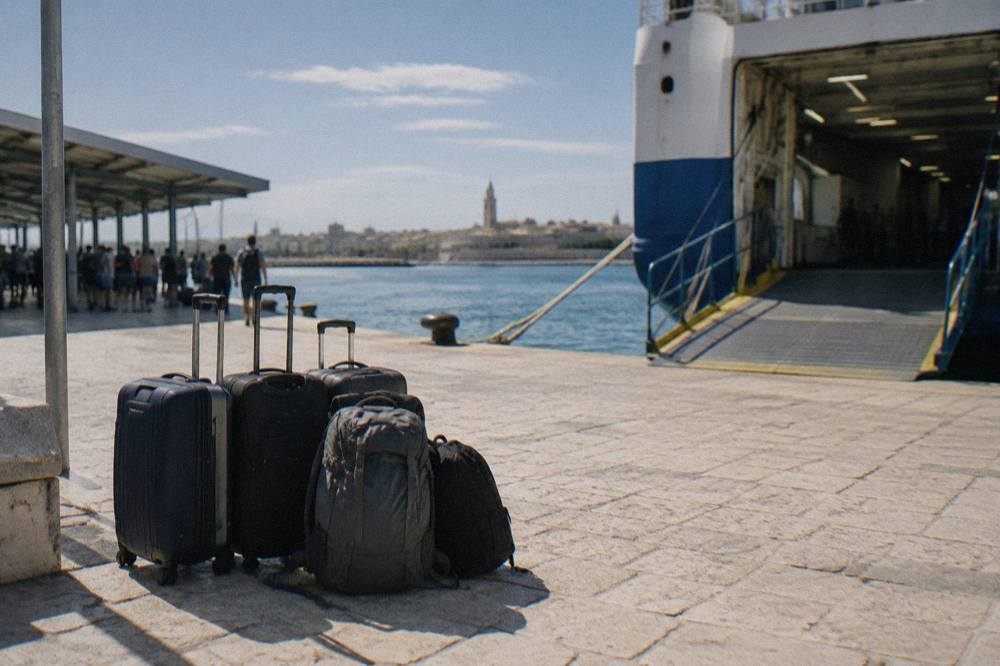 Suitcases and rucksacks stacked on a sunlit quay beside a ferry gangway with travellers waiting in the shade