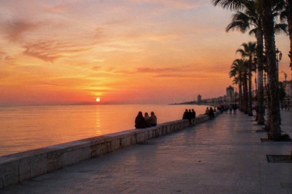 Seafront promenade at sunset with distant people on the low sea wall, an orange sky and palm silhouettes