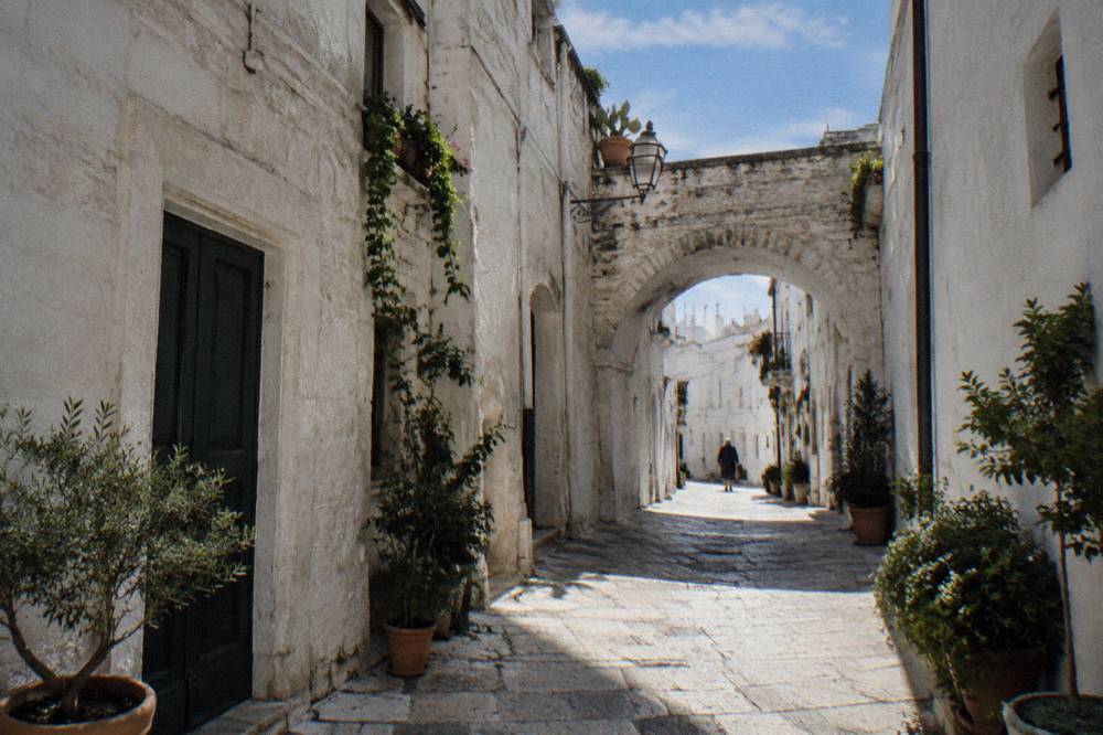 Narrow white old town lane with a stone arch and potted plants in afternoon shade