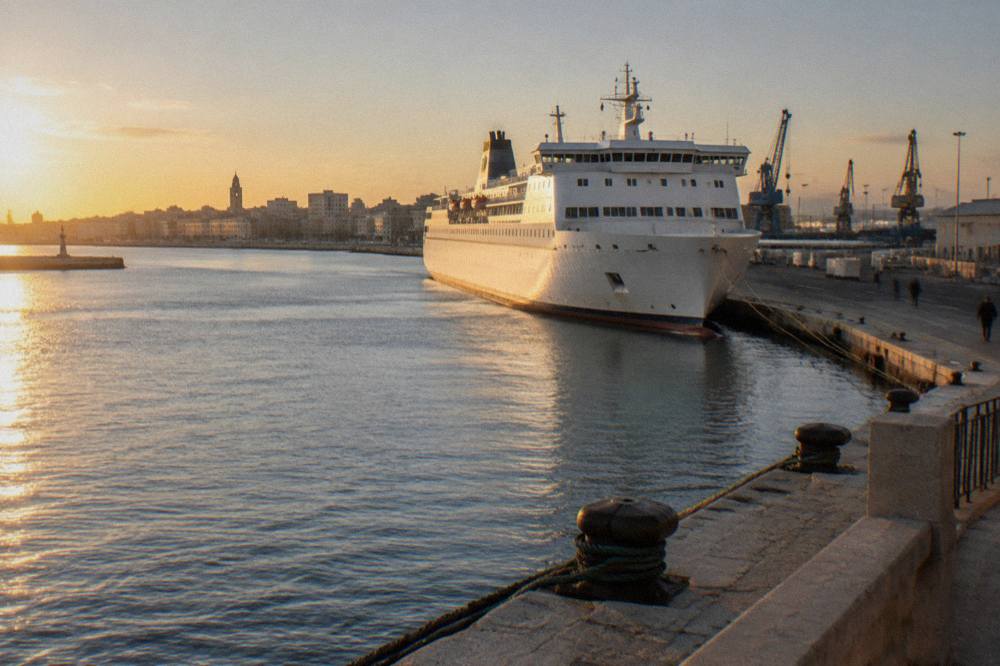 Large white passenger ferry moored at a long quay with bollards and ropes and cranes beyond