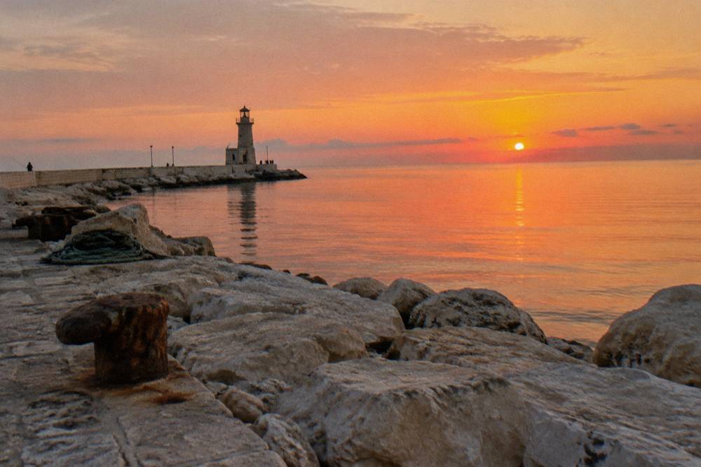 End of a harbour pier with a small lighthouse, glassy open Adriatic and a distant angler at sunset