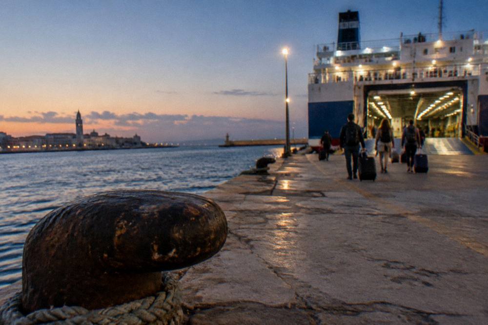 Distant passengers with wheeled suitcases walking along a quay toward a ferry's open loading ramp in evening light