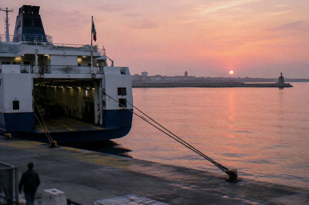 Stern of a ferry at a quay at sunrise with a pink sky, taut mooring ropes and still water