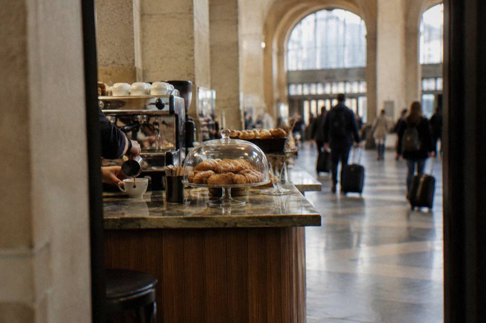 Station cafe counter with hands pouring an espresso and pastries under a glass dome