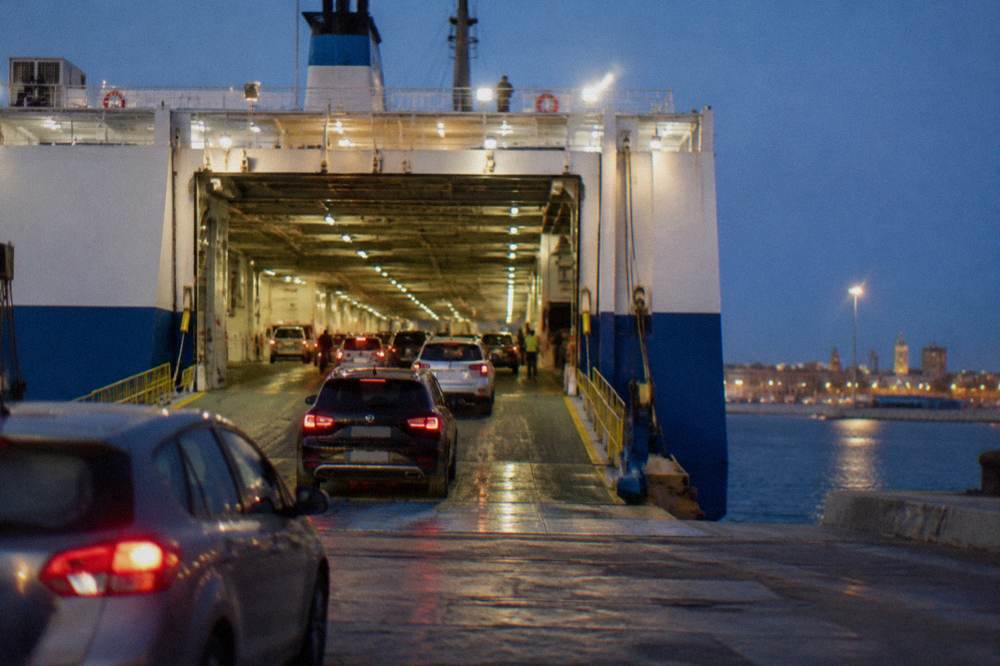 Ferry vehicle deck seen from the quay with rows of cars driving in under floodlights at evening