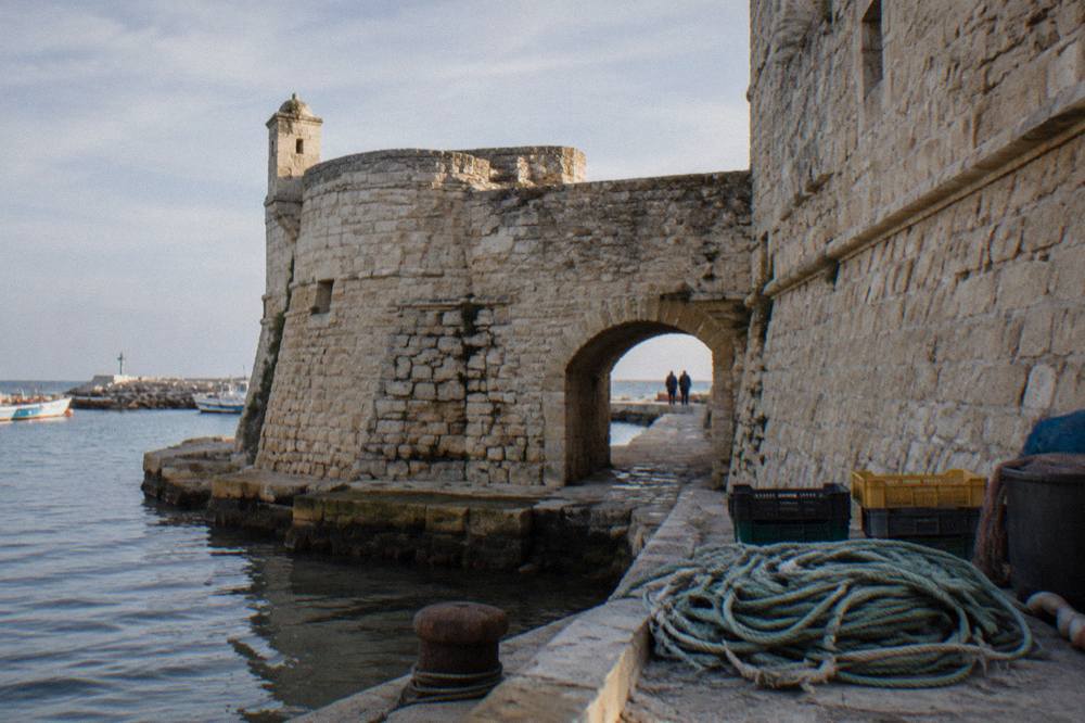 Weathered limestone fort walls and a low arched gateway beside harbour water with coiled ropes and crates on the quay