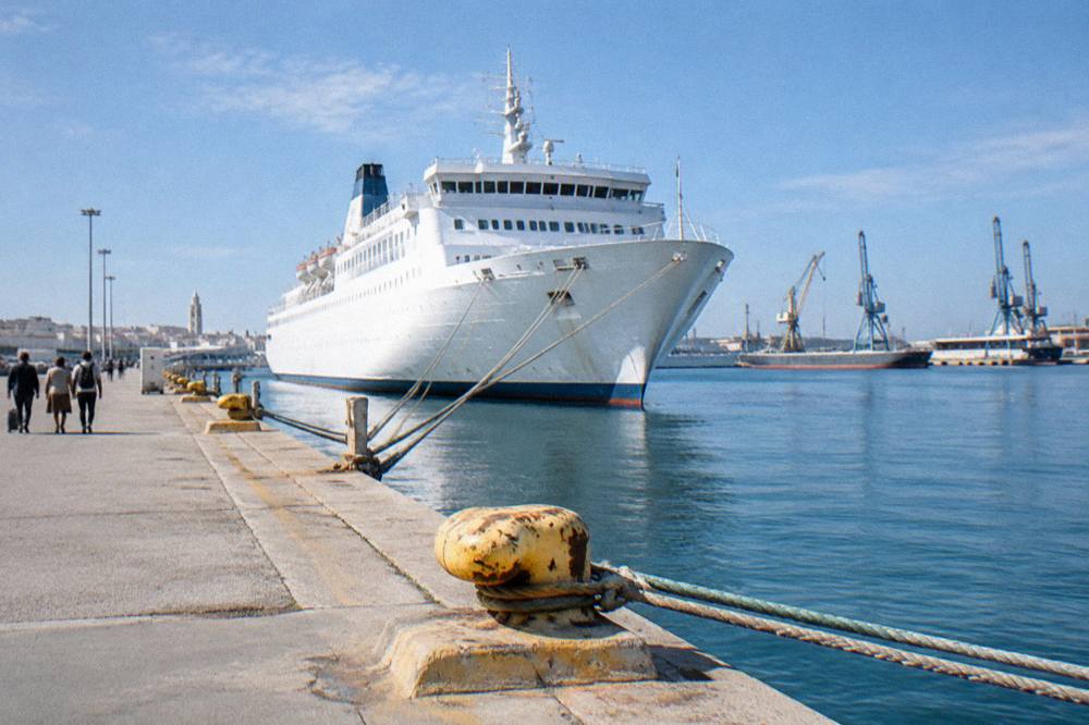 Large white passenger ferry moored at a long quay with bollards and ropes and cranes beyond