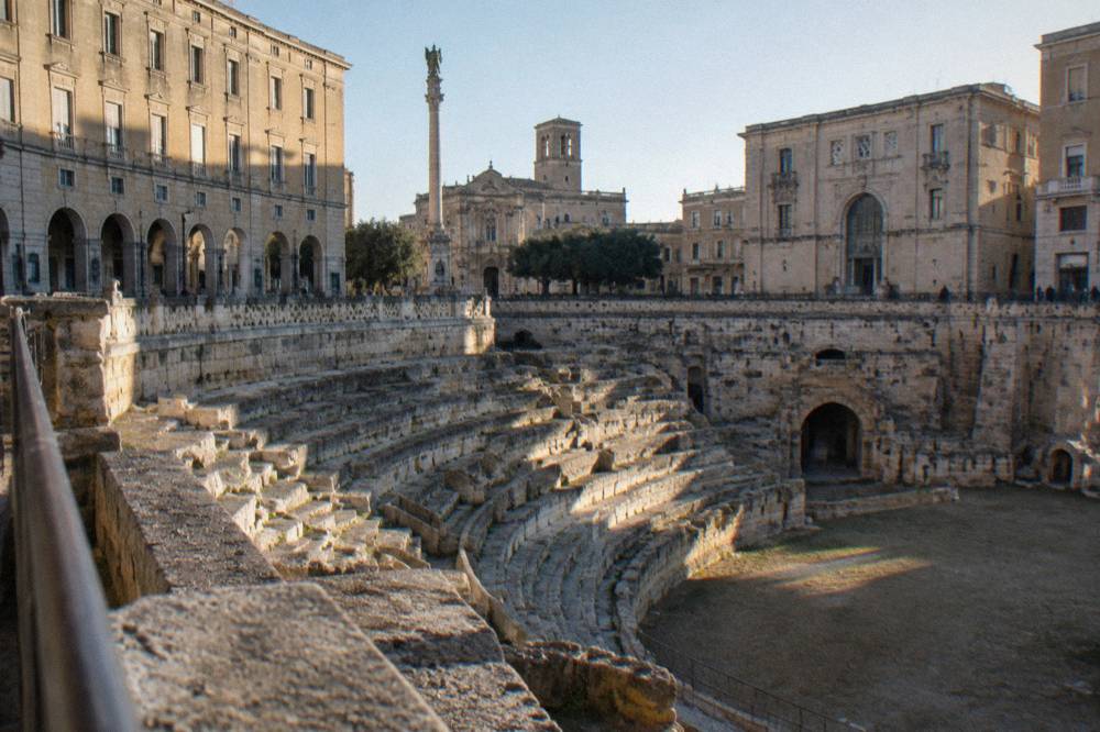 Roman amphitheatre half excavated in a city square surrounded by palazzi in afternoon light