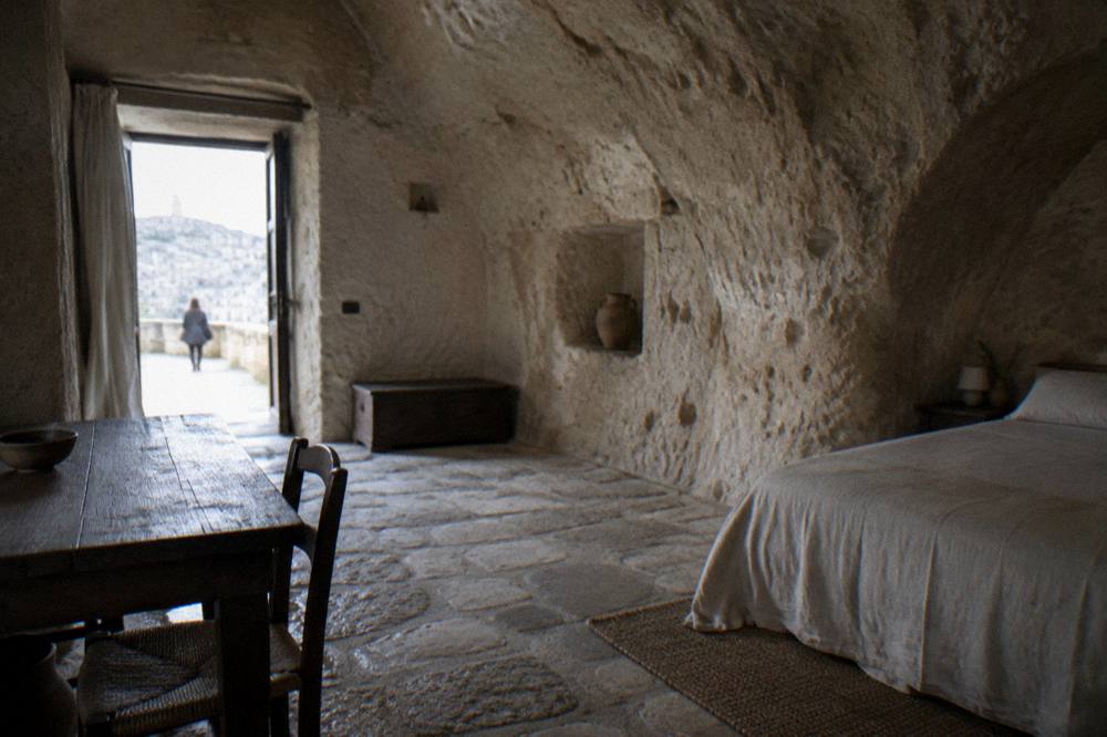 Inside a restored cave dwelling with an arched rock ceiling, a simple bed and a wooden table