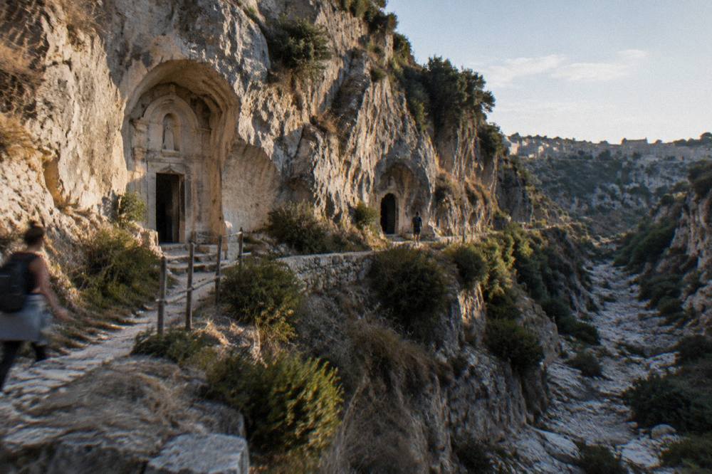 Cave entrances cut into a pale rock face above a dry stream bed with shrubs on the ledges and a distant hiker