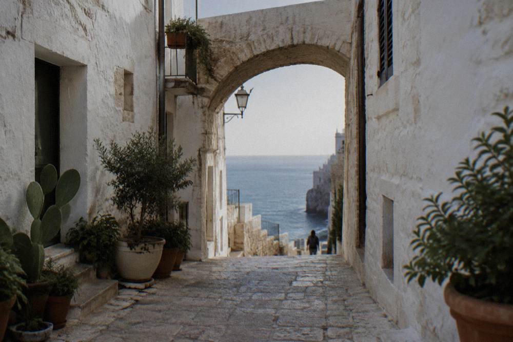 Narrow whitewashed cliff-top old town lane with a stone arch, potted plants and a glimpse of blue sea