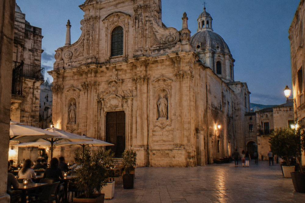 Baroque cathedral facade in warm pale stone on a small square with a dome behind and cafe tables