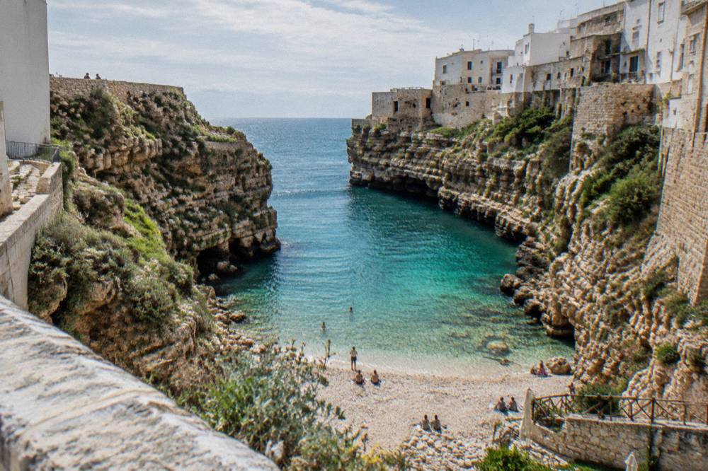 View down from a stone bridge into a narrow cove of clear turquoise water and white pebbles