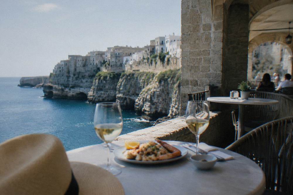 Small round tables on a cliff-edge terrace with glasses and a plate of seafood above blue water