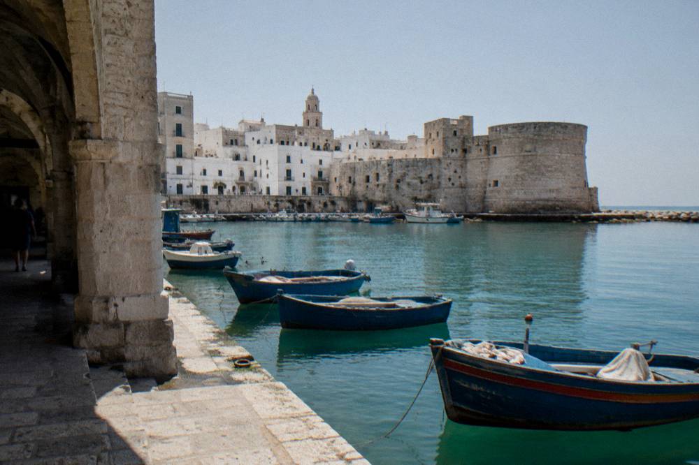 Old harbour of small painted wooden boats with a stone castle on the headland and a whitewashed old town behind