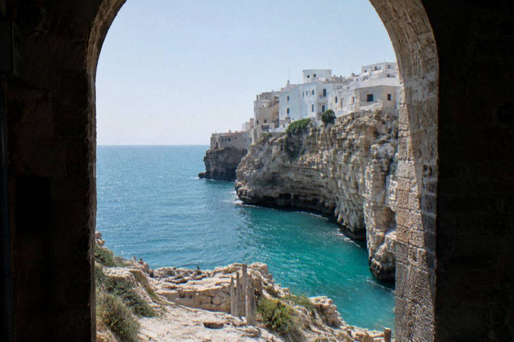 White town perched on the edge of tall pale limestone cliffs above a turquoise sea seen from a rocky viewpoint