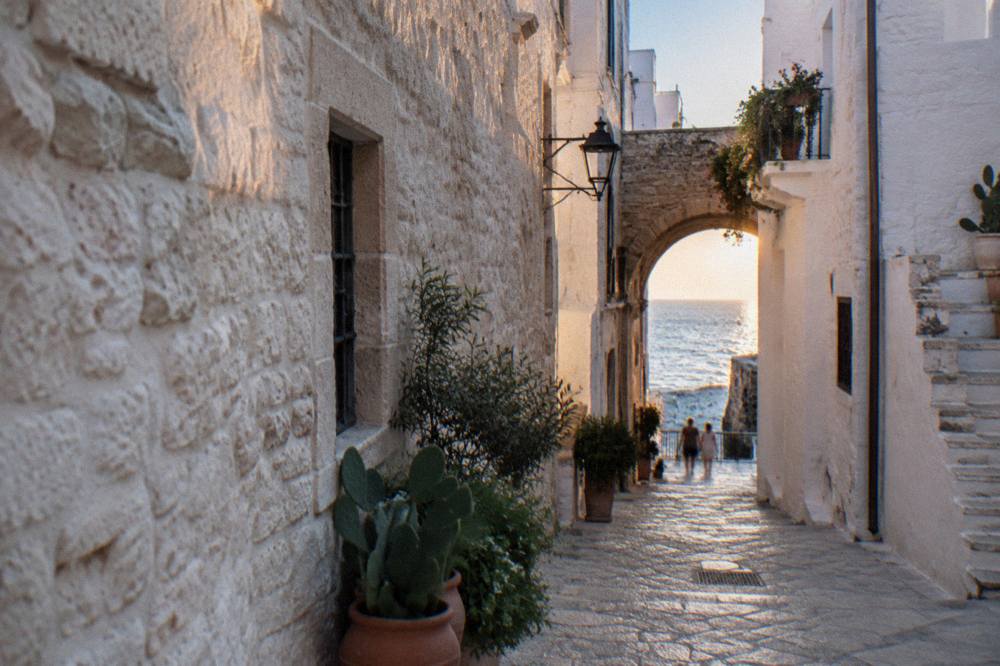Narrow whitewashed cliff-top old town lane with a stone arch, potted plants and a glimpse of blue sea