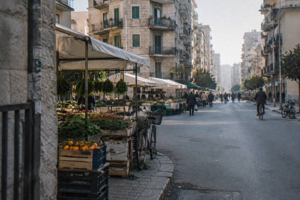 Residential street of apartment blocks with fruit and vegetable stalls under plain awnings and distant shoppers